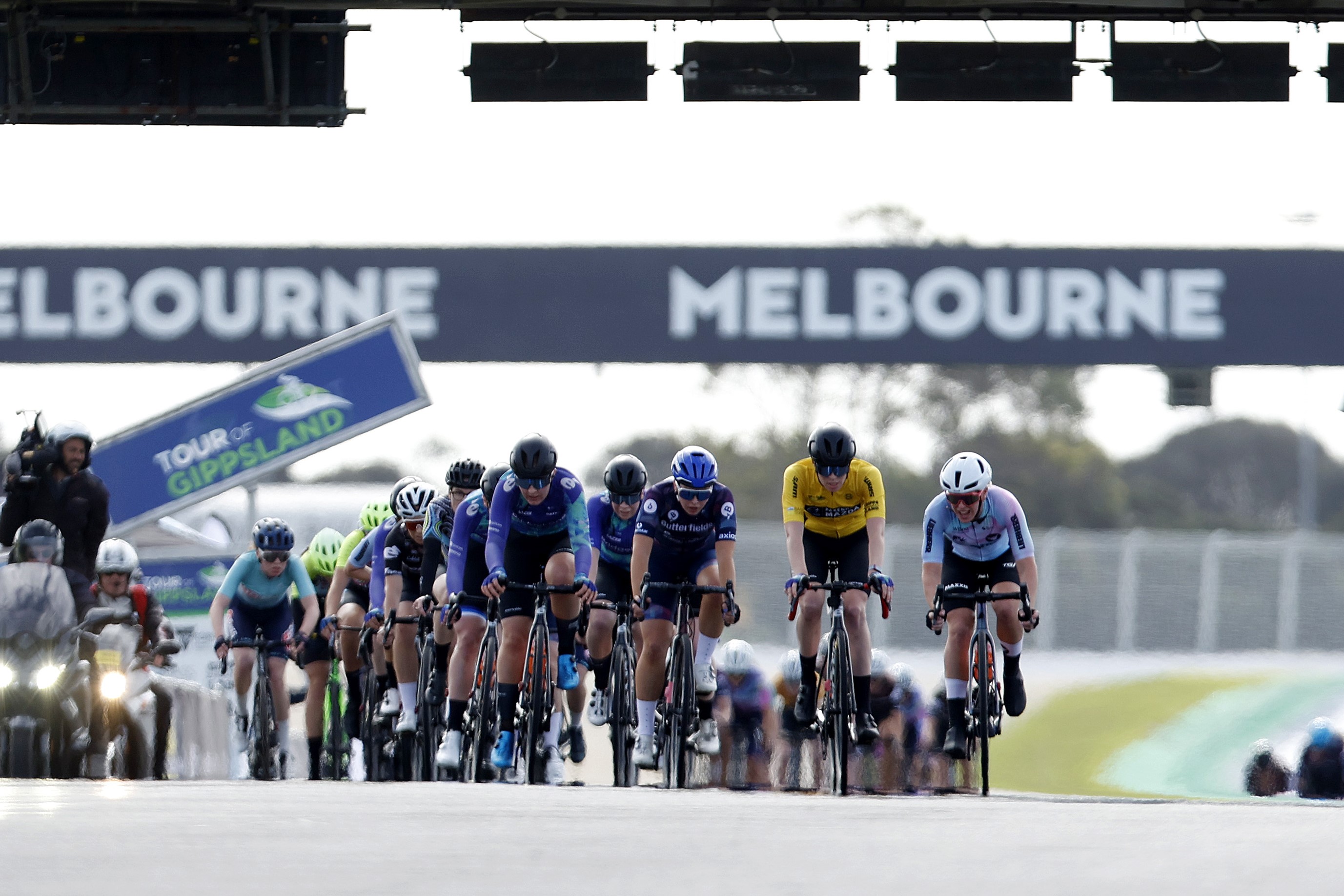 Peloton in the 2023 Tour of Gippsland women's road race on the Phillip Island Grand Prix motor racing circuit, with a banner saying 'Melbourne' in the background. Photo by Con Chronis.